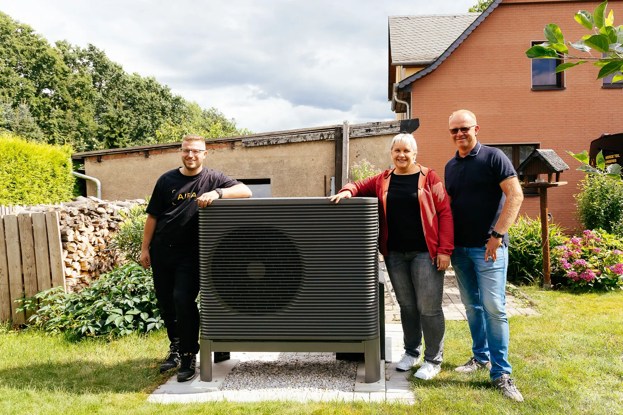 Ein Ehepaar plus ein Wärmepumpen-Installateur stehen im Garten an einem Wärmepumpen-Außengerät