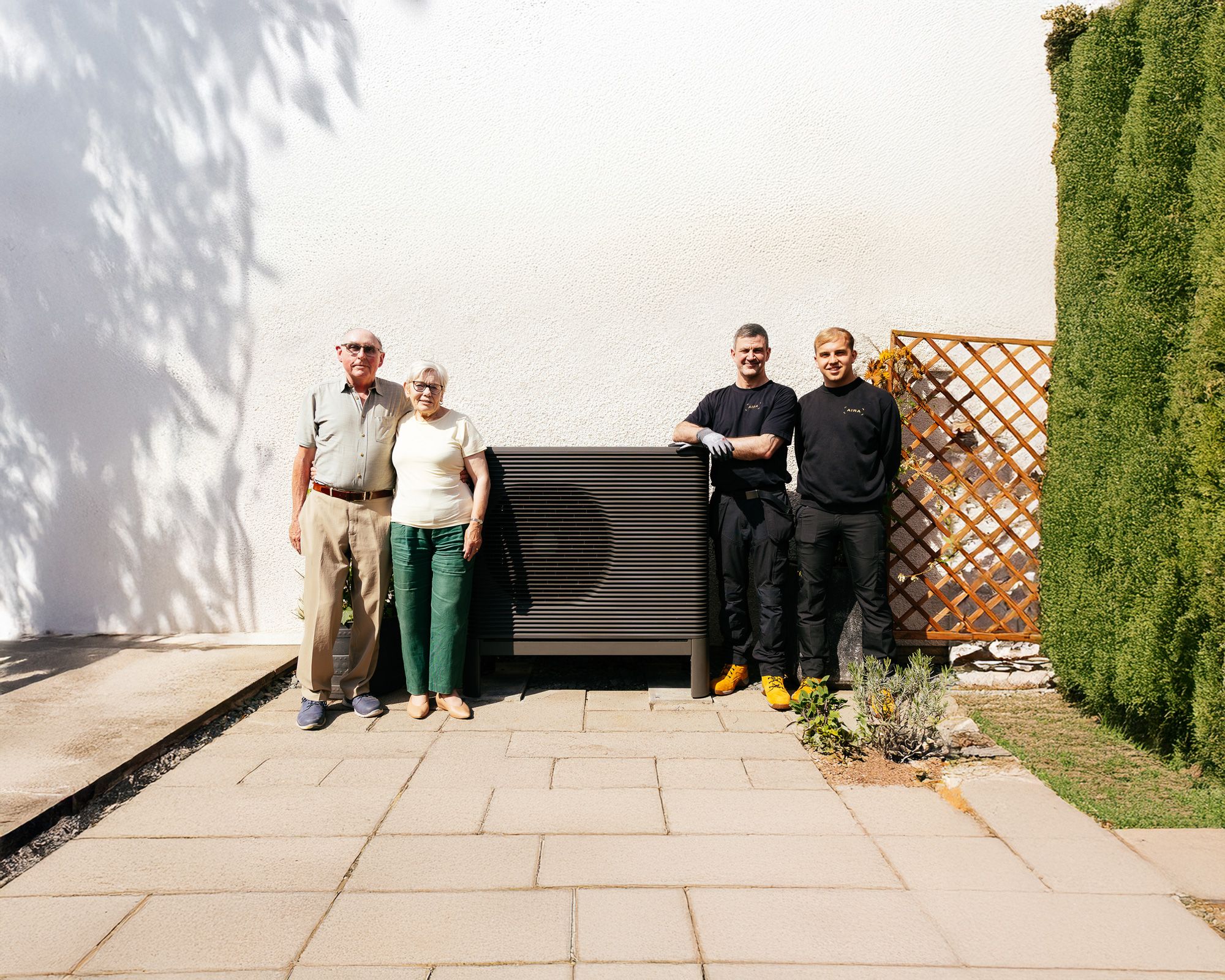 Ein Paar steht mit zwei Installateuren vor Ihrer installierten Wärmepumpe, welche an einer weißen Hauswand platziert ist