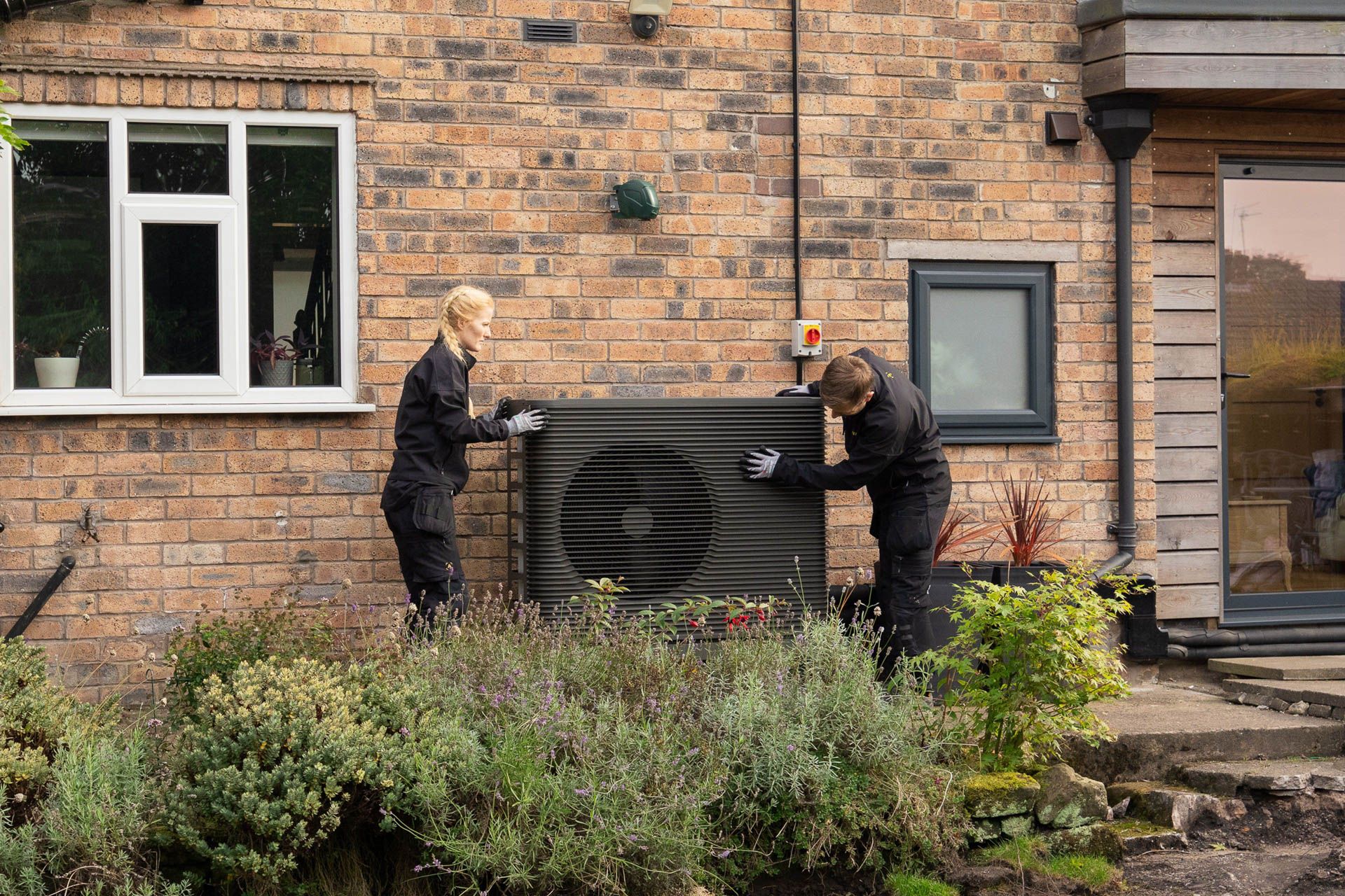 Zwei Installateure von Aira stehen neben einer Wärmepumpe vor einer weißen Wand im Garten