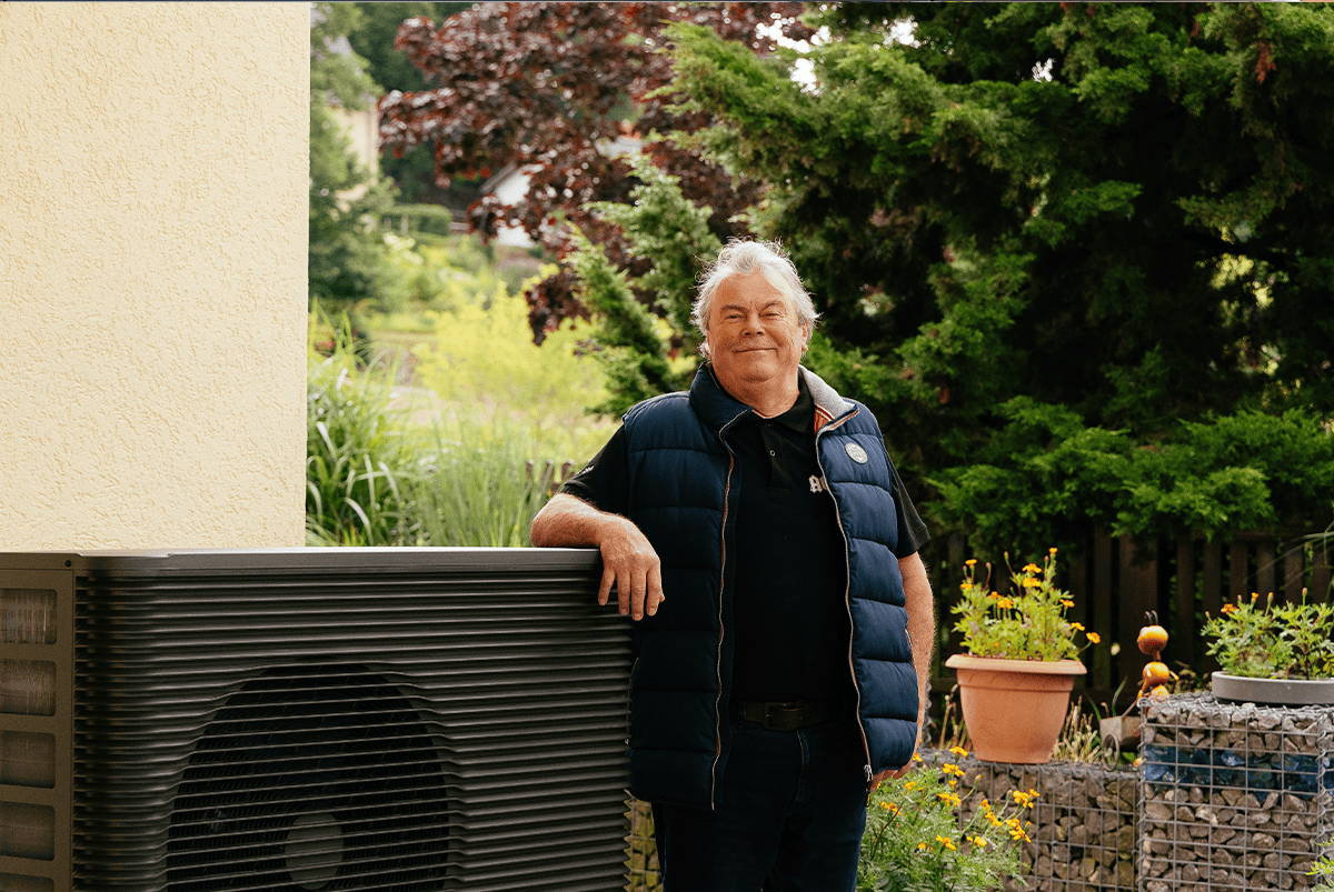Eine zufriedene Person steht neben seiner Wärmepumpe im Garten vor der Hauswand